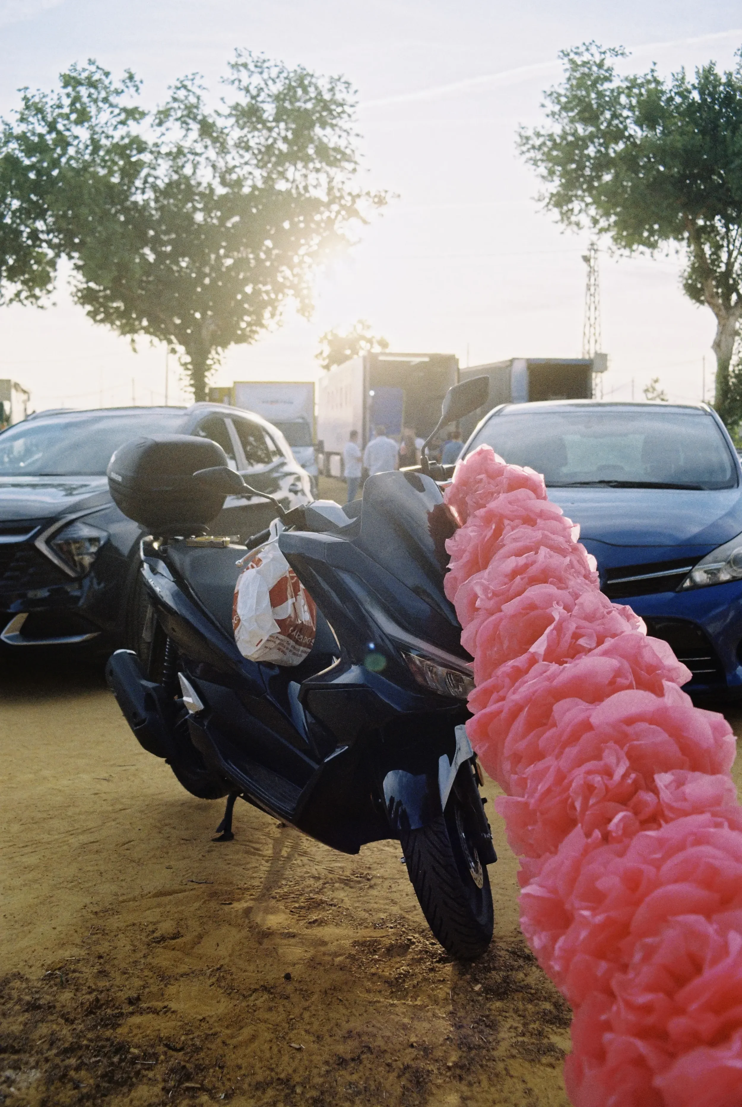 Estacionamiento al aire libre con varios autos y una motocicleta negra decorada con una cadena de globos rosas, al atardecer con árboles y personas al fondo.