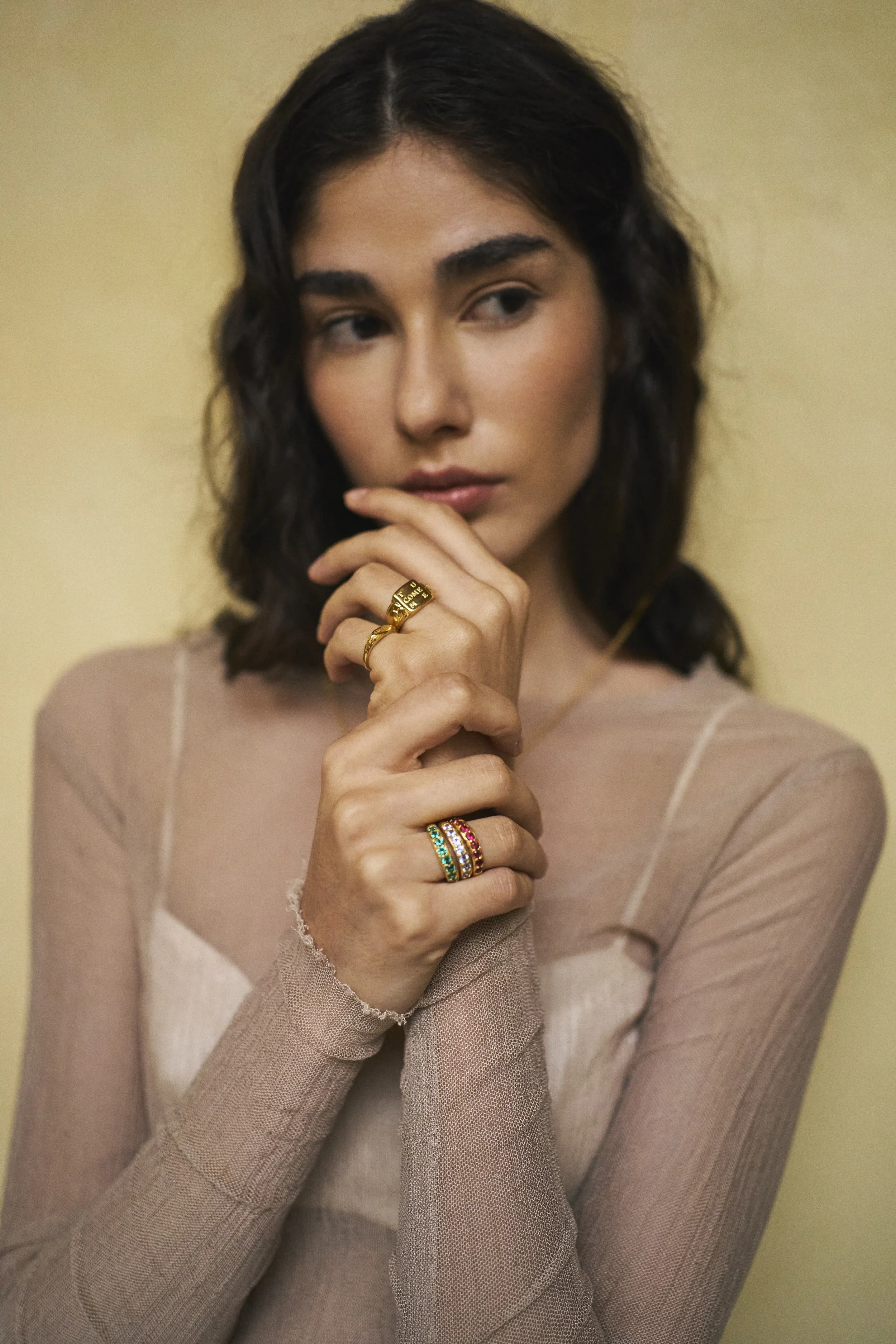 Retrato de una mujer joven con cabello rizado oscuro, que lleva varias pulseras y anillos en las manos, con un fondo de pared color crema.