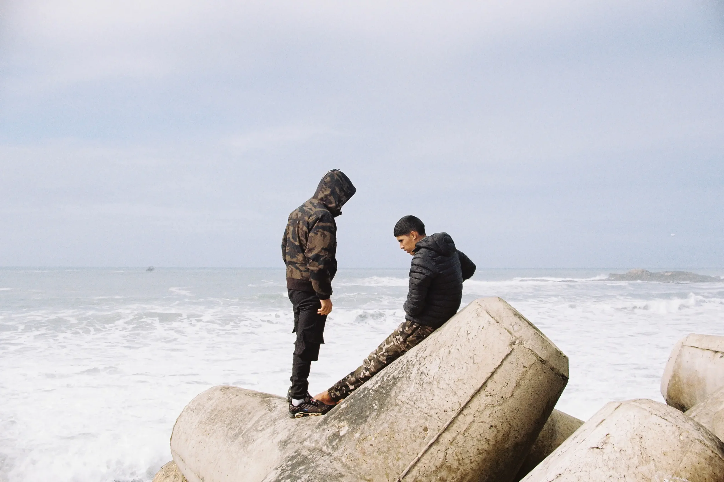 Dos niños en la playa sobre bloques de cemento. Uno está de pie y el otro sentado, ambos con ropa de abrigo y mirando al suelo, con un mar y cielo despejado de fondo.