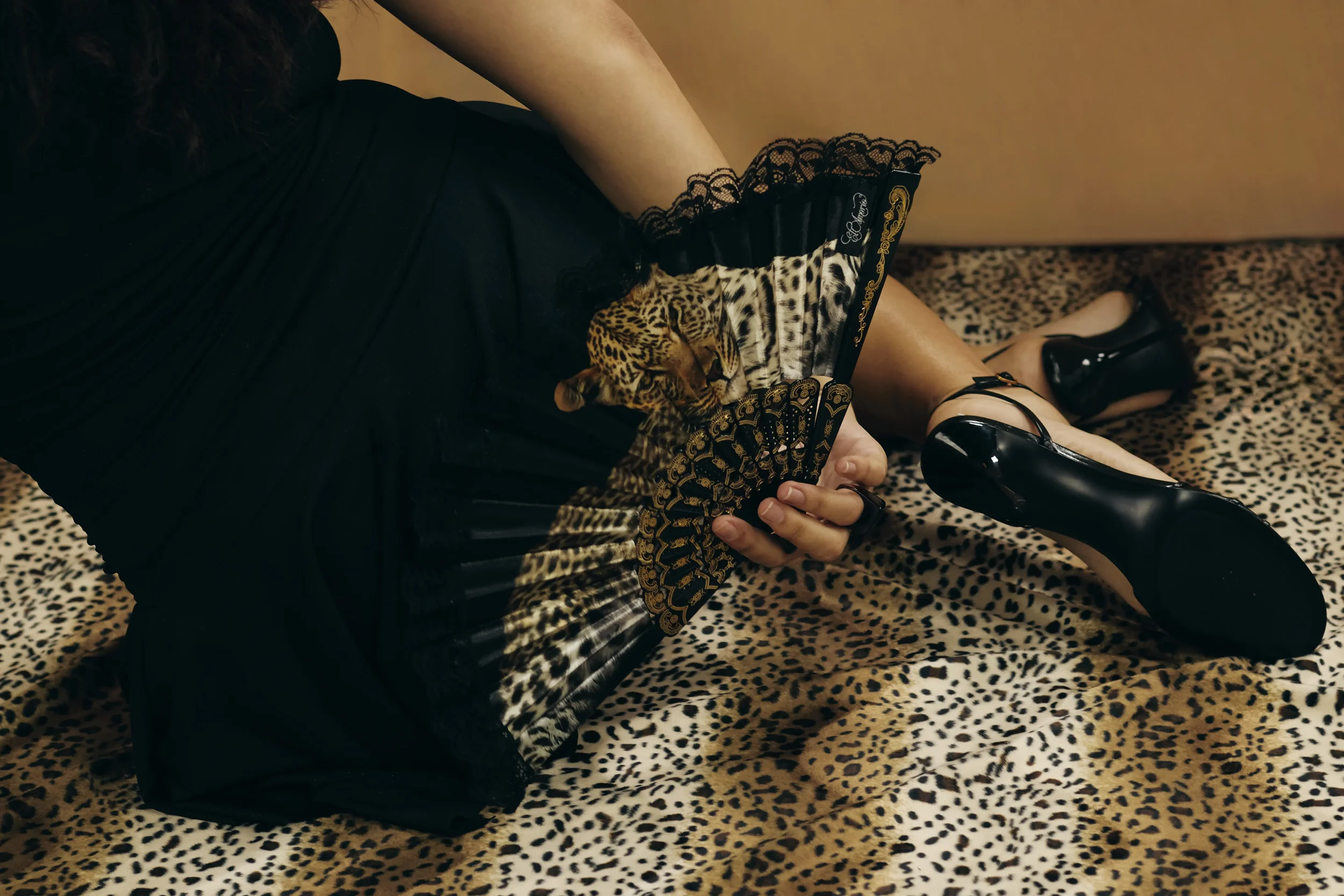 Persona con vestido negro y tacones negros sentado sobre una alfombra de estampado leopardo, sosteniendo una revista con la imagen de un jaguar, en un fondo de pared beige.