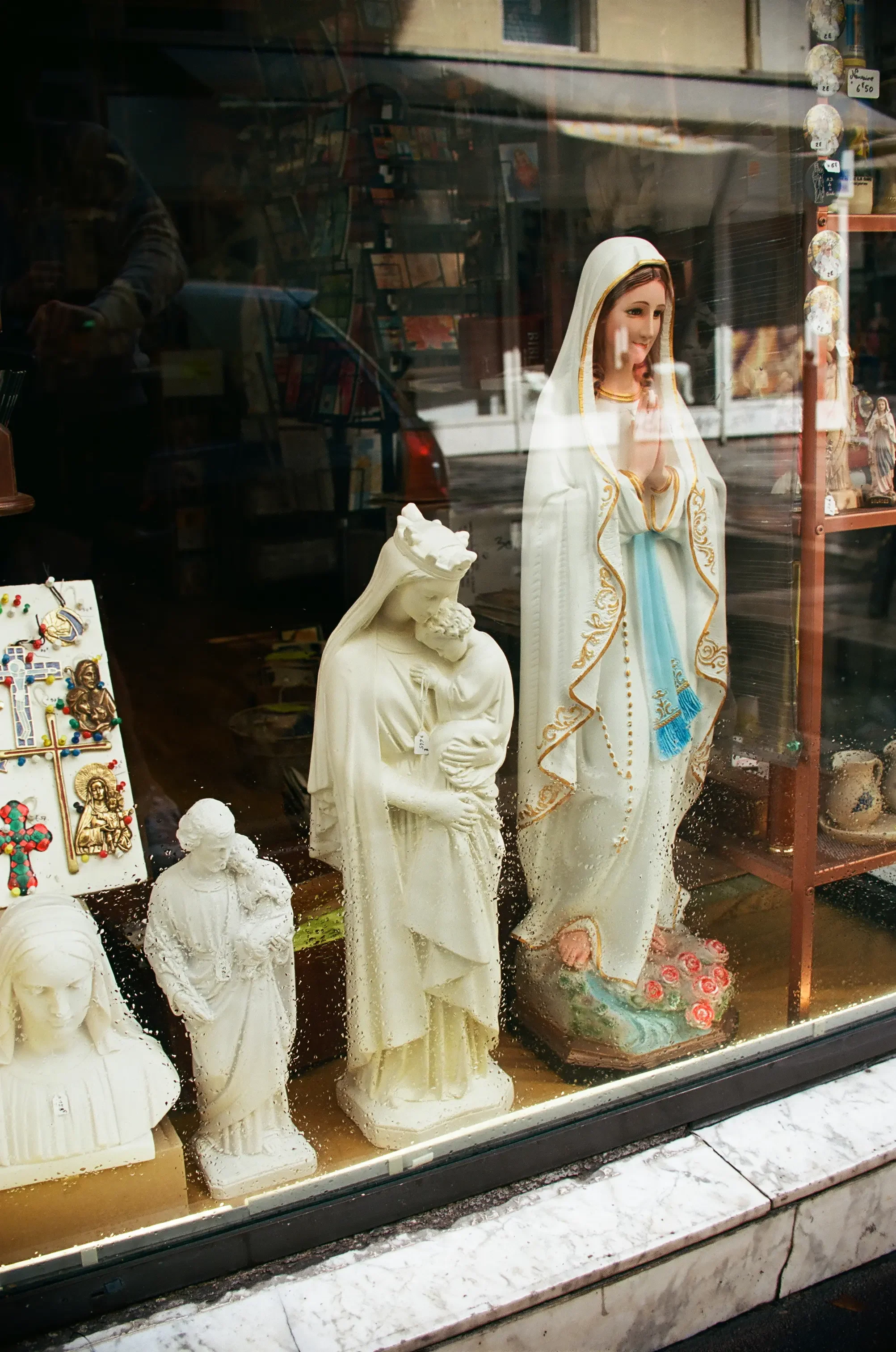 Figuras de yeso de la Virgen María y varios santos en una tienda, con el reflejo de la calle en la ventana.