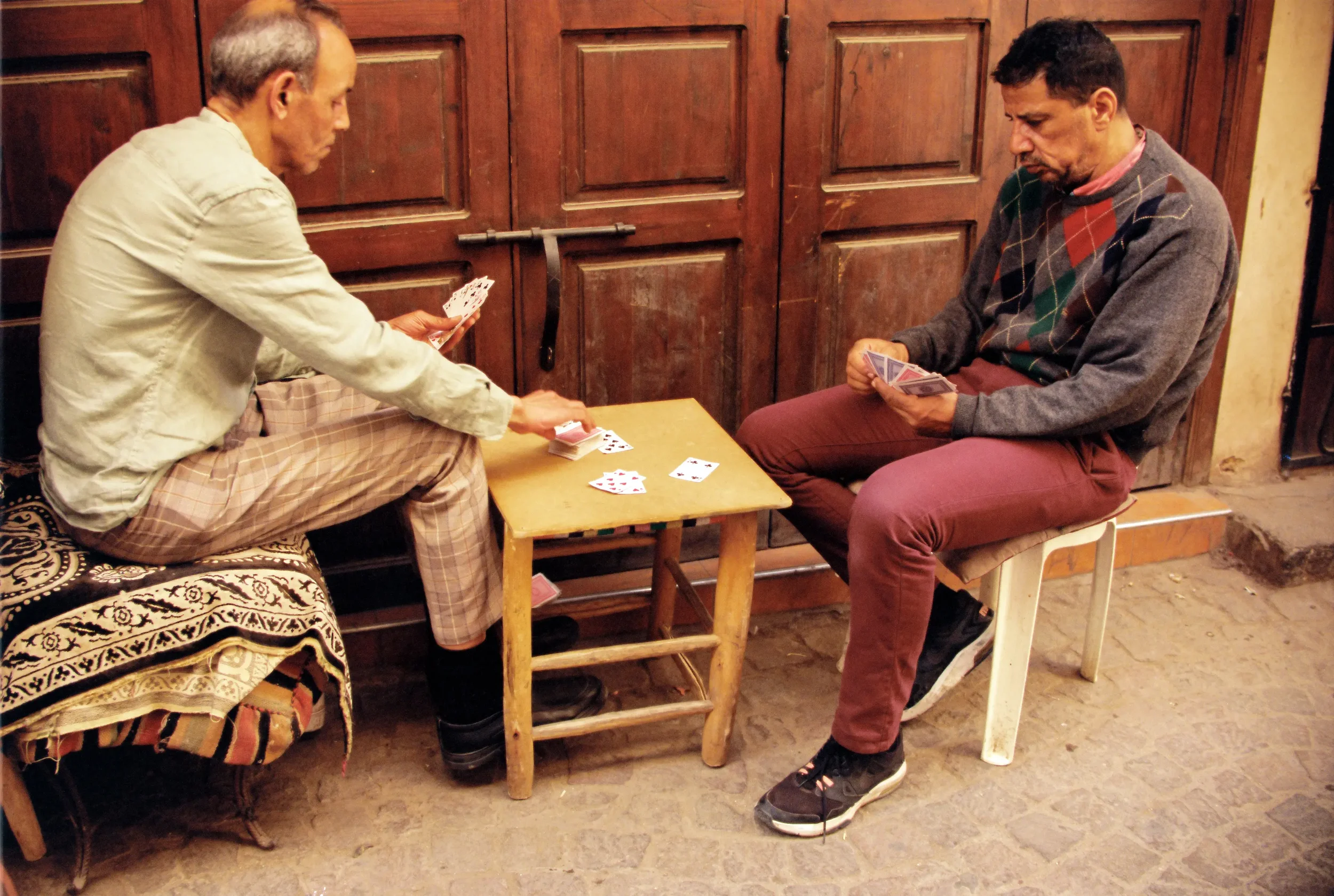 Dos hombres sentados en una mesa jugando cartas, uno con un suéter a cuadros y el otro con una chaqueta beige, en un ambiente con paredes de madera.