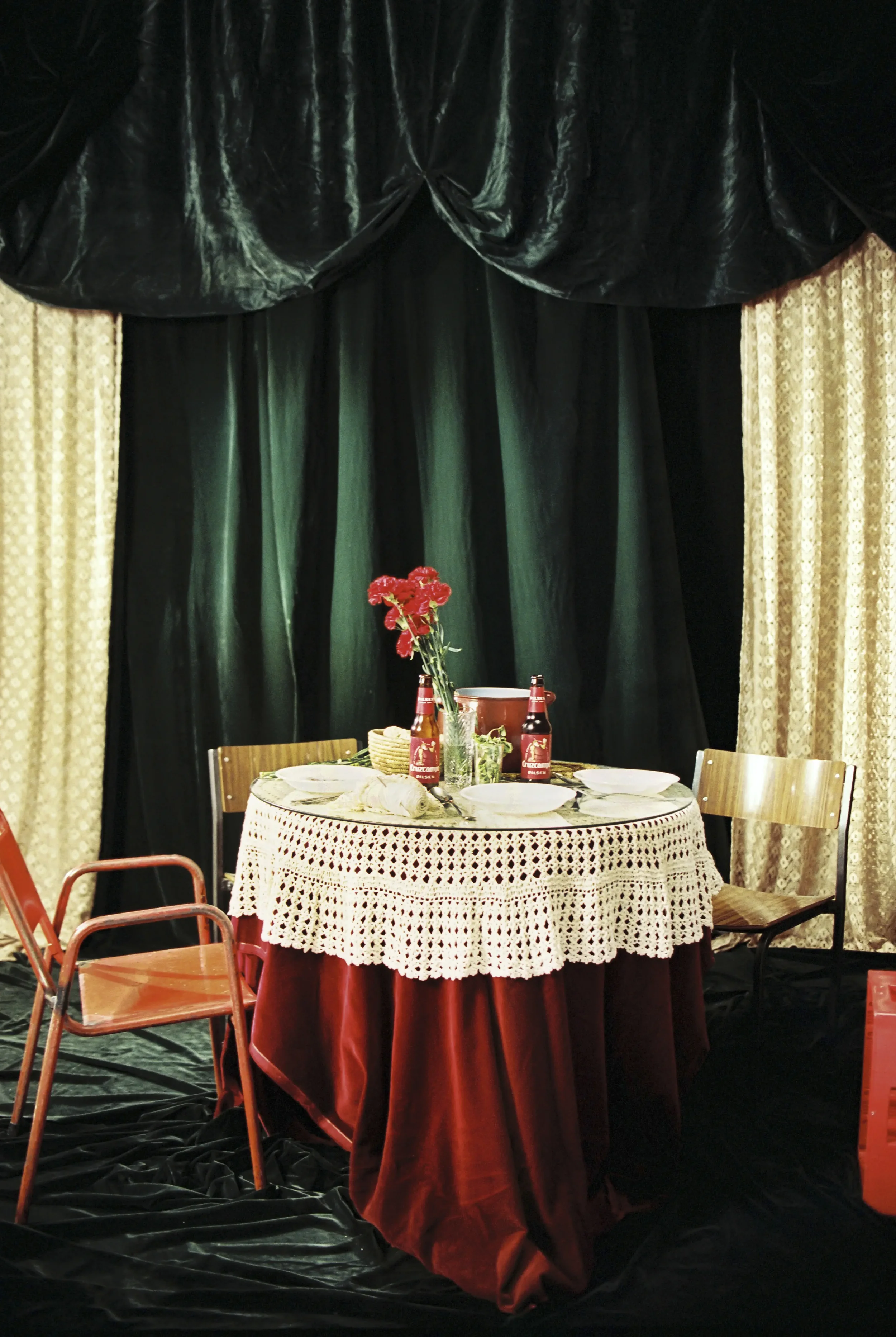 Mesa decorada con un mantel de encaje, flores rojas en un florero, comida, y dos botellas de salsa de tomate en un entorno con cortinas y fondo oscuro.
