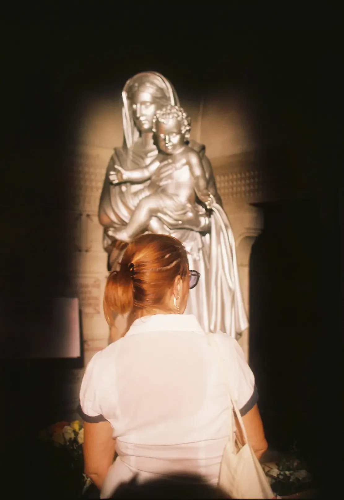 Mujer con cabello al hombro, vista desde atrás, observa una estatua de María con Niño Jesús en un museo o iglesia.