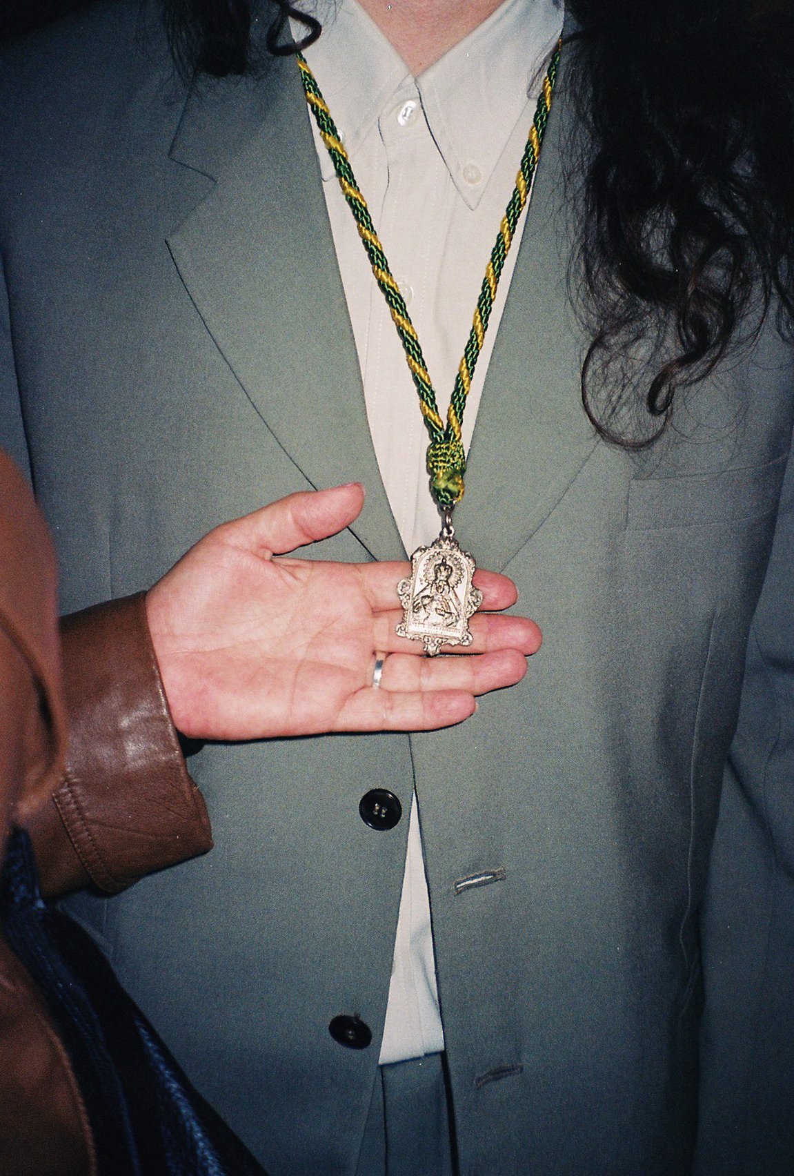 Persona usando saco gris claro, camisa blanca y collar de hilo amarillo, verde y negro con medalla o relicario colgando.