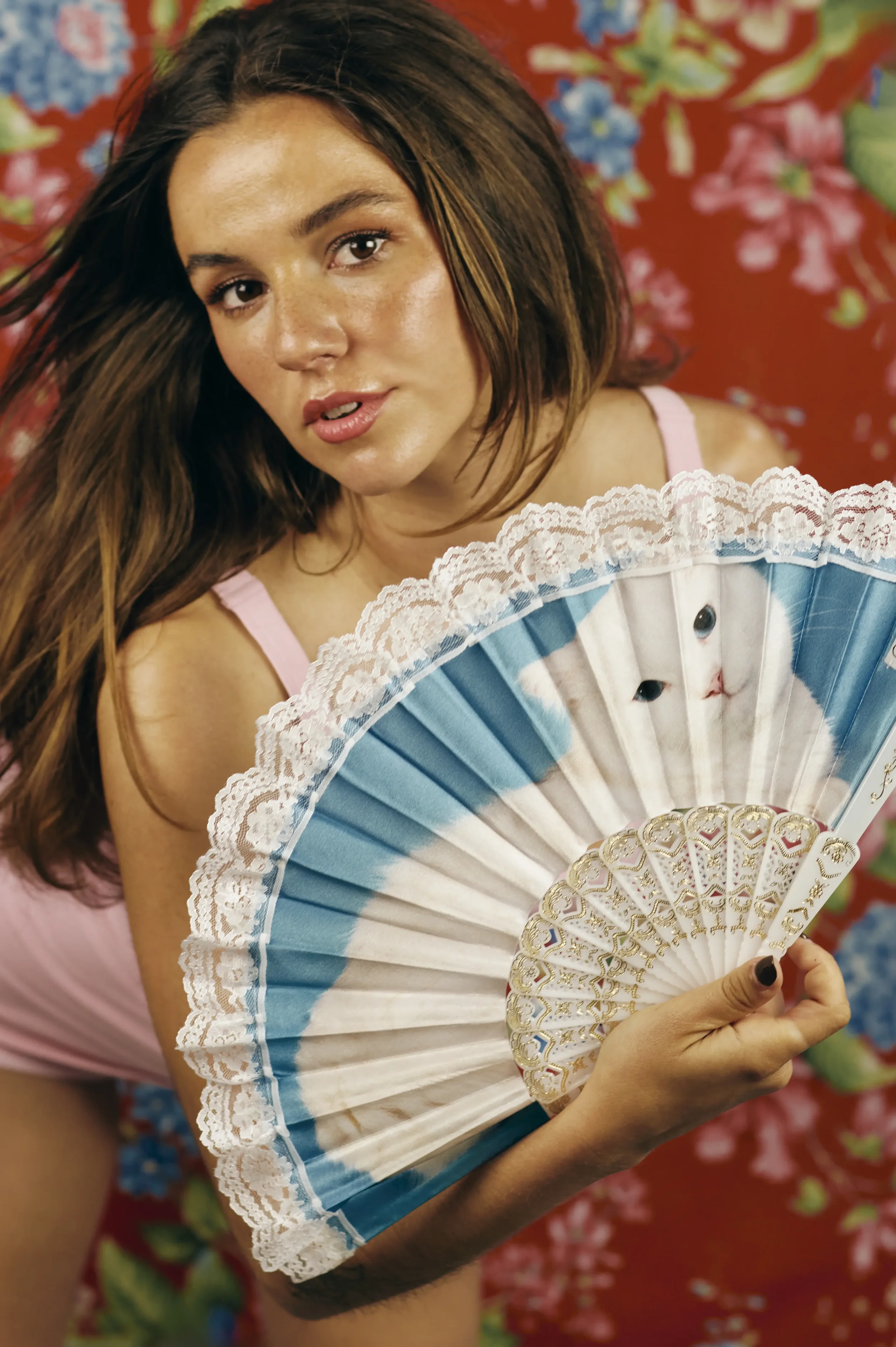 Mujer con cabello largo y peinado suelto, usando blusa rosa, sostiene un abanico decorativo con detalles dorados y encaje, en un fondo con patrón floral de colores vibrantes.