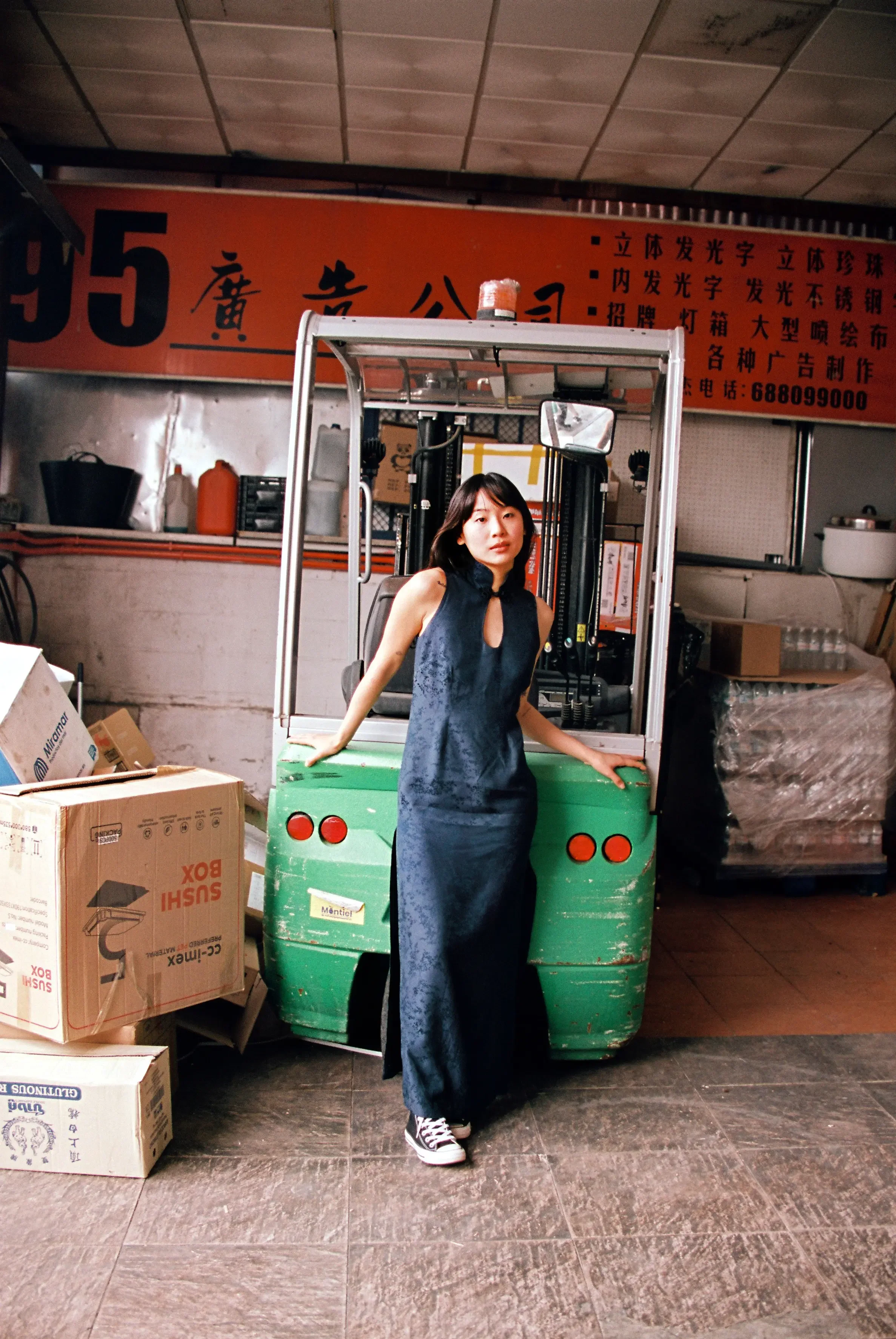 Mujer con vestido largo y zapatillas deportivas, de pie junto a un montacargas en un almacén o tienda, con cajas y estantes con productos alrededor, y un cartel naranja con caracteres en idioma chino en el fondo.