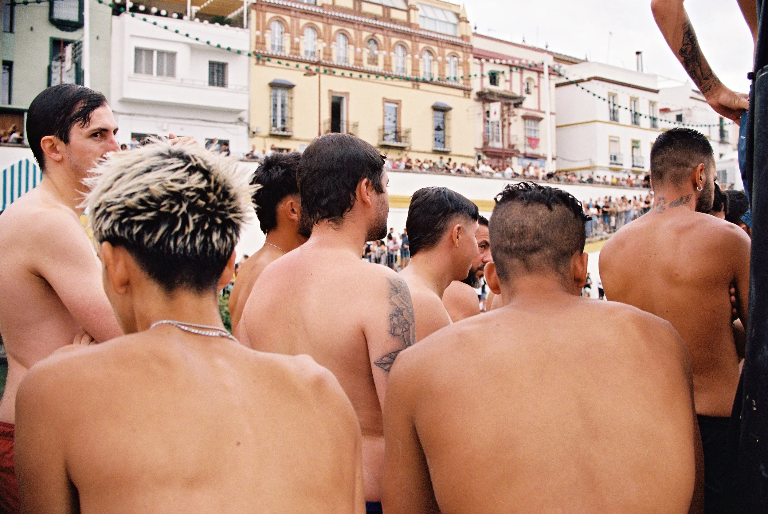 Grupo de personas sin camiseta en una plaza pública, con edificios antiguos y una multitud al fondo.