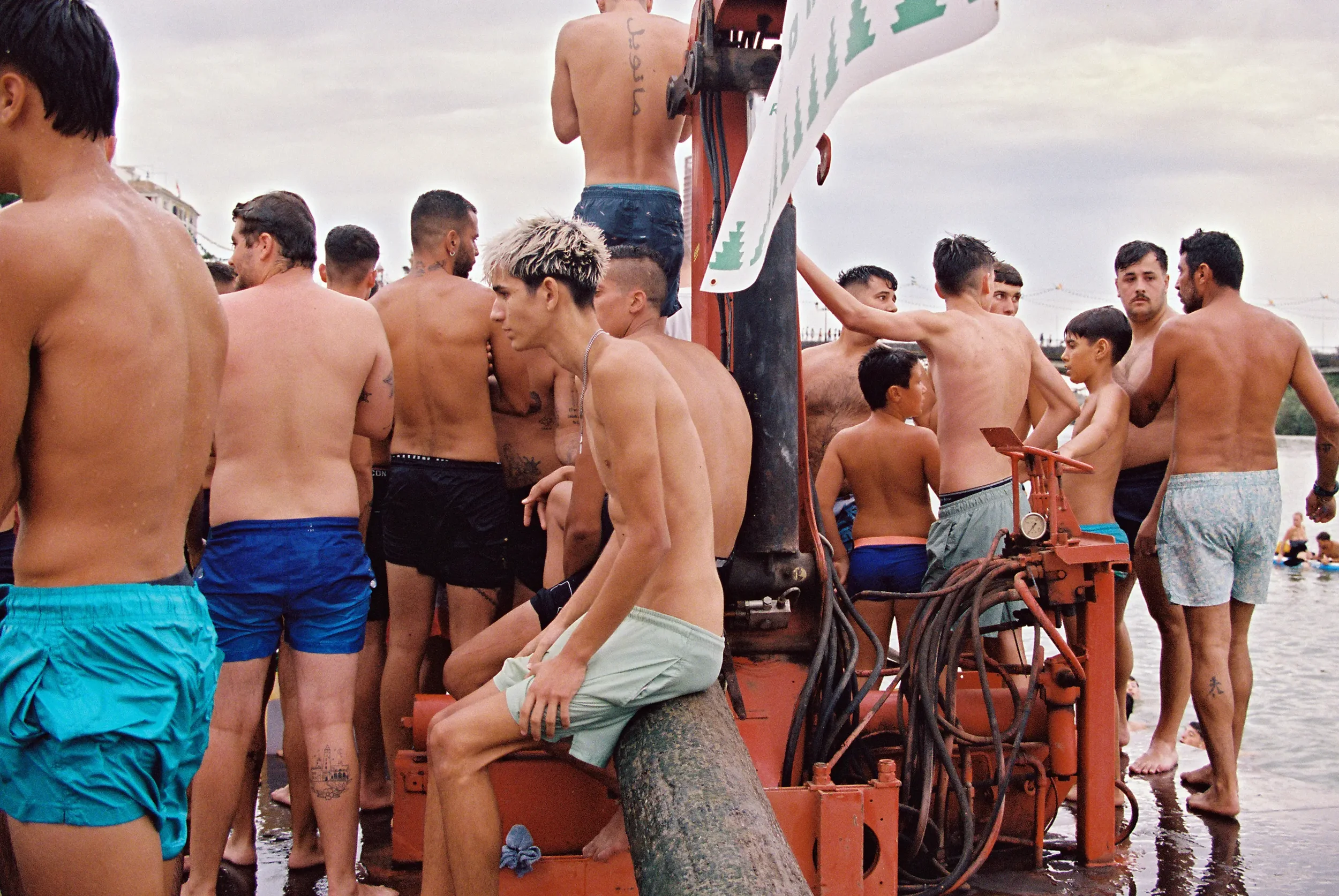 Grupo de hombres, algunos sin camisa, en una playa participando en una actividad de natación o pesca. Hay accesorios y equipo náutico visible y el cielo parece nublado.