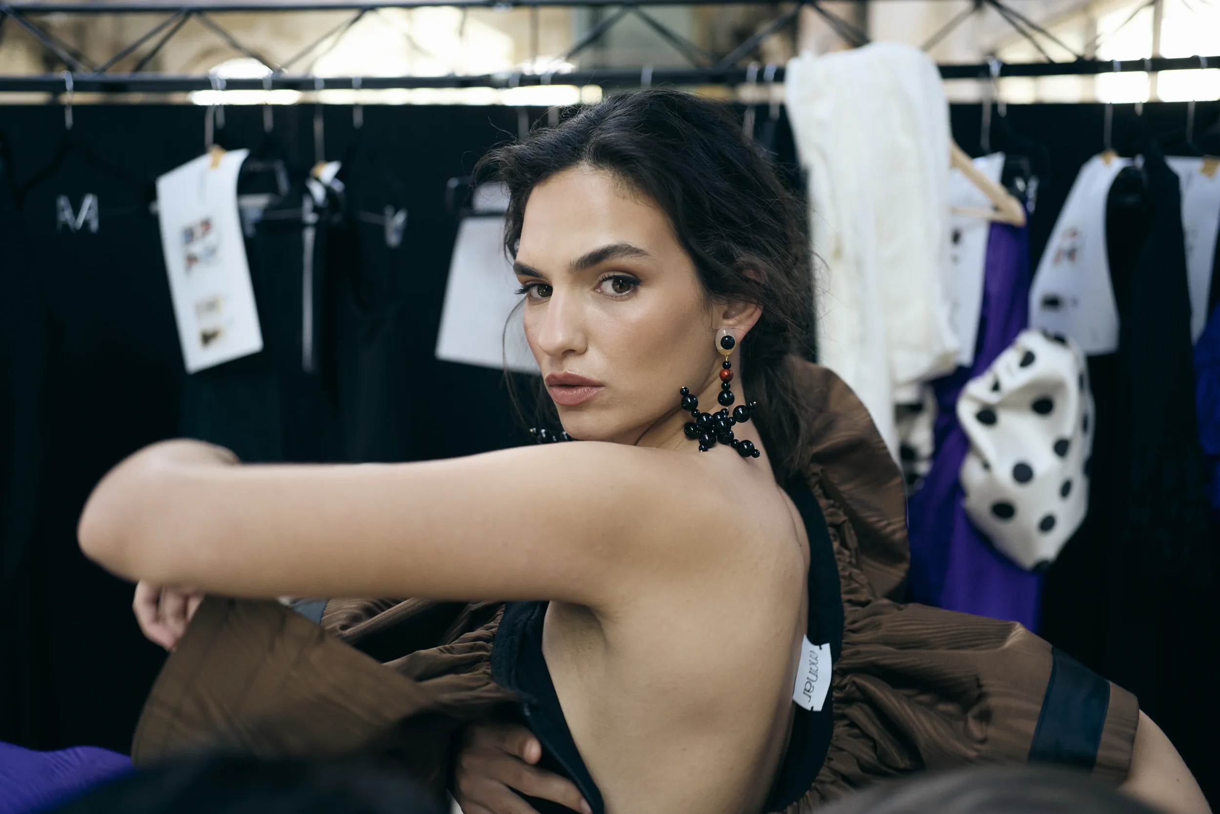 Mujer rubia con pendientes grandes de perlas negras y bolas rojas, en un fondo de ropa colgada en una percha, en un ambiente de vestidor o tienda de ropa.