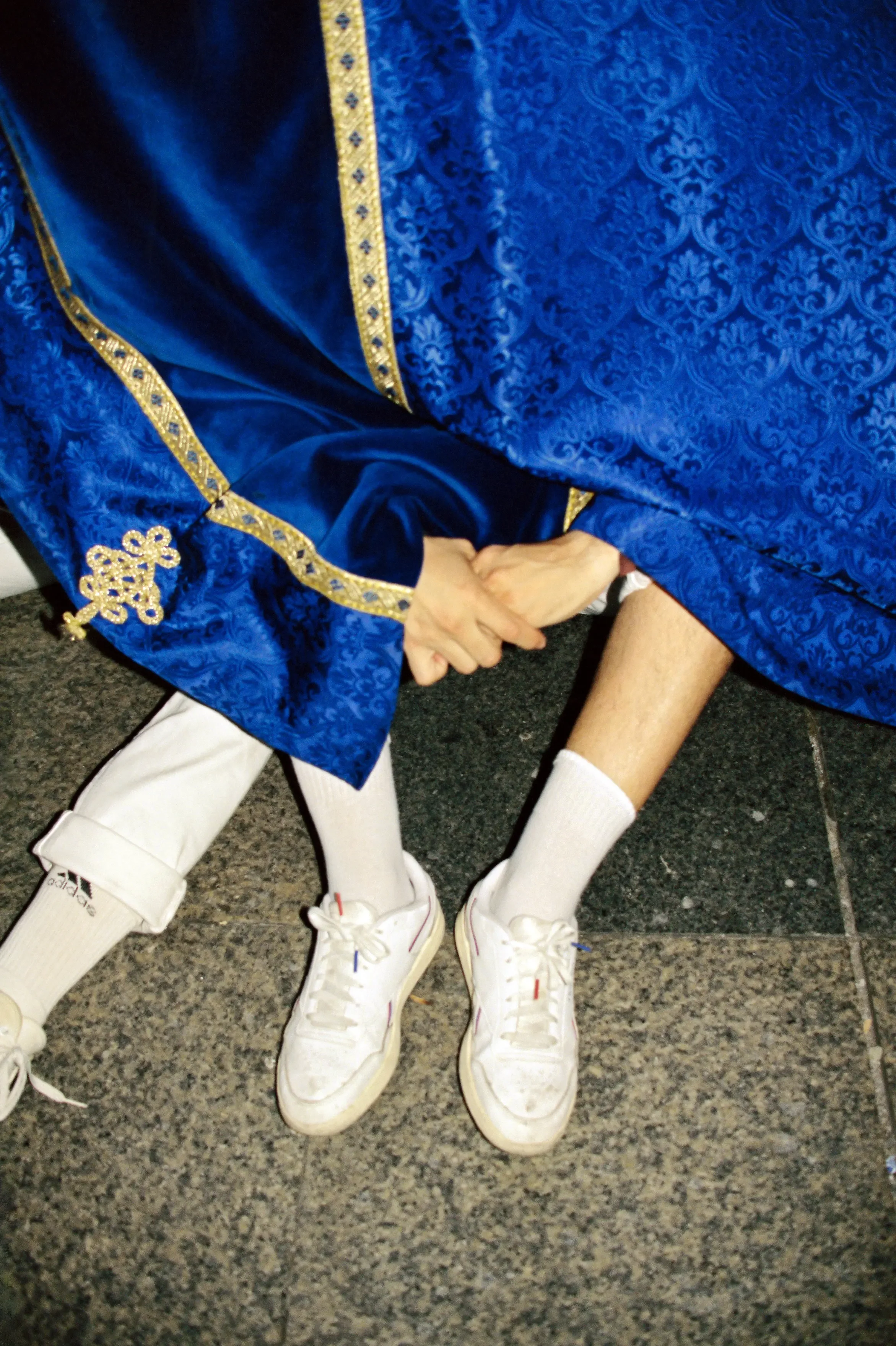 Dos niños con uniforme escolar, usan zapatillas blancas, calcetas largas blancas, y se toman de las manos con un adulto vestido con toga azul con detalles dorados, en un piso de piedra.