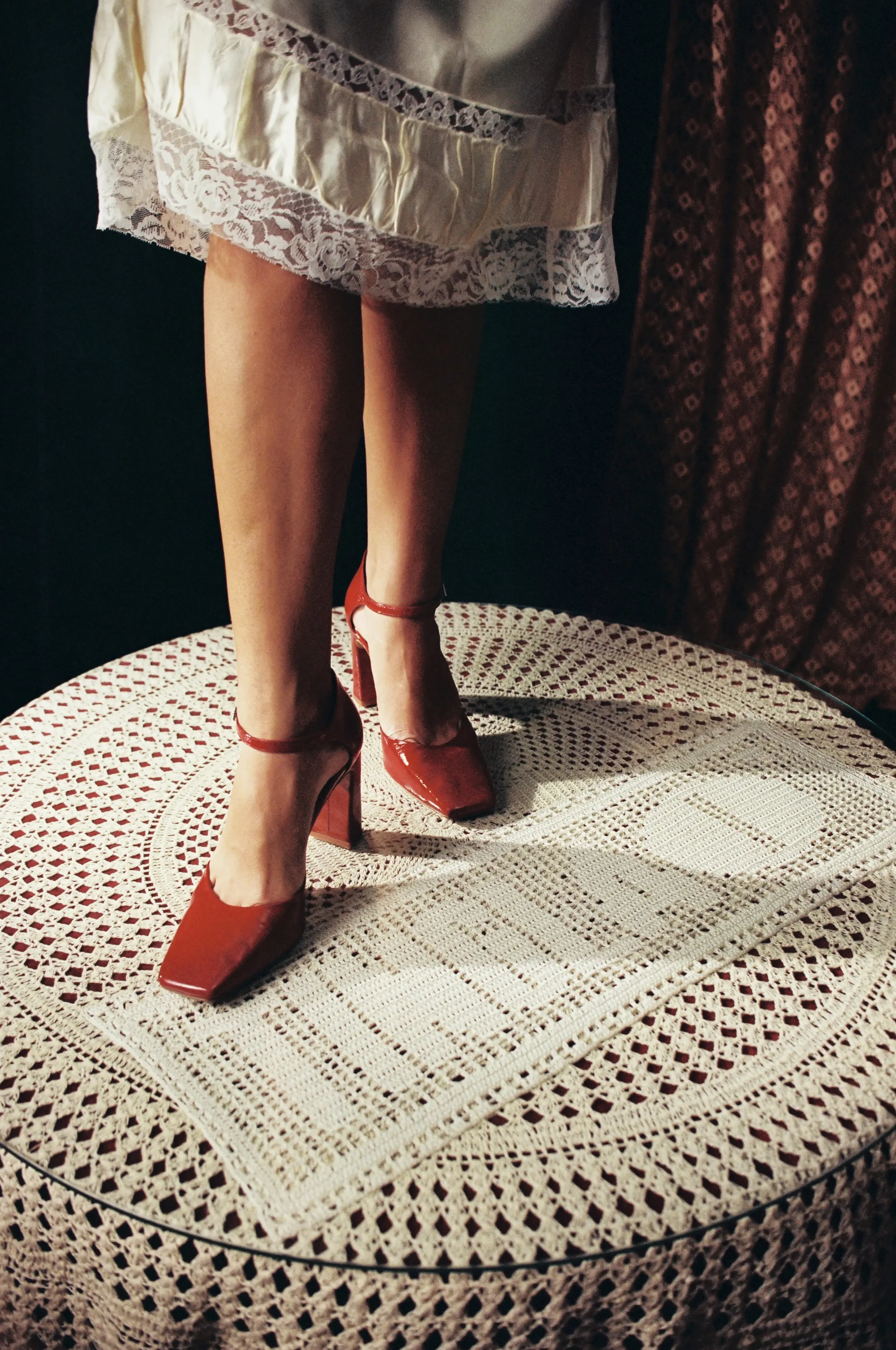 Piernas femeninas con tacones rojos, falda de encaje y una mesa de crochet en primer plano. Fondo con cortinas oscuras con patrón floral.
