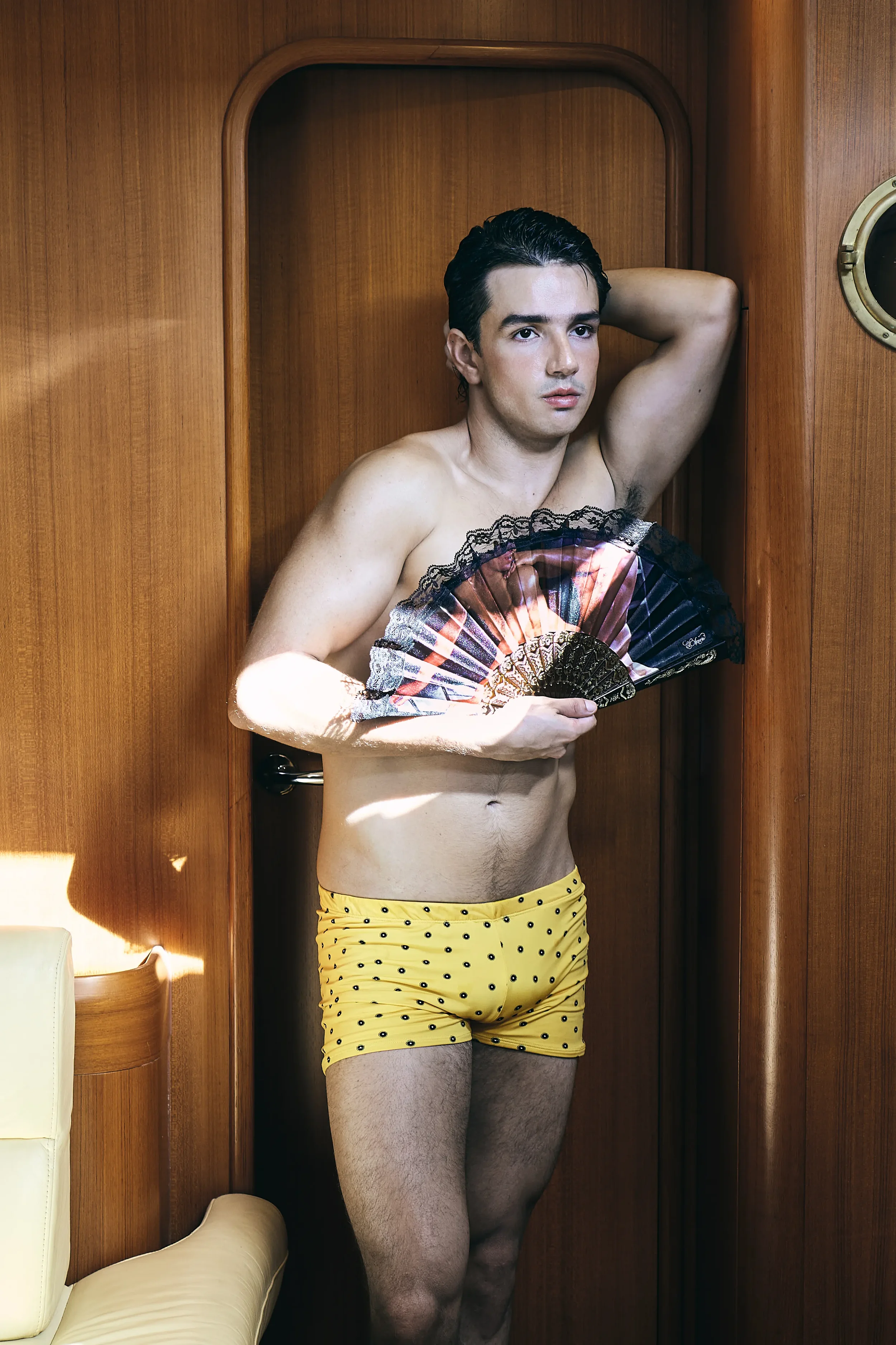 Hombre rubio con bigote, sin camiseta, con pantalones cortos amarillos con puntos negros, sosteniendo un abanico, de pie en una cabina de madera, con una mano detrás de la cabeza y la otra con abanico.
