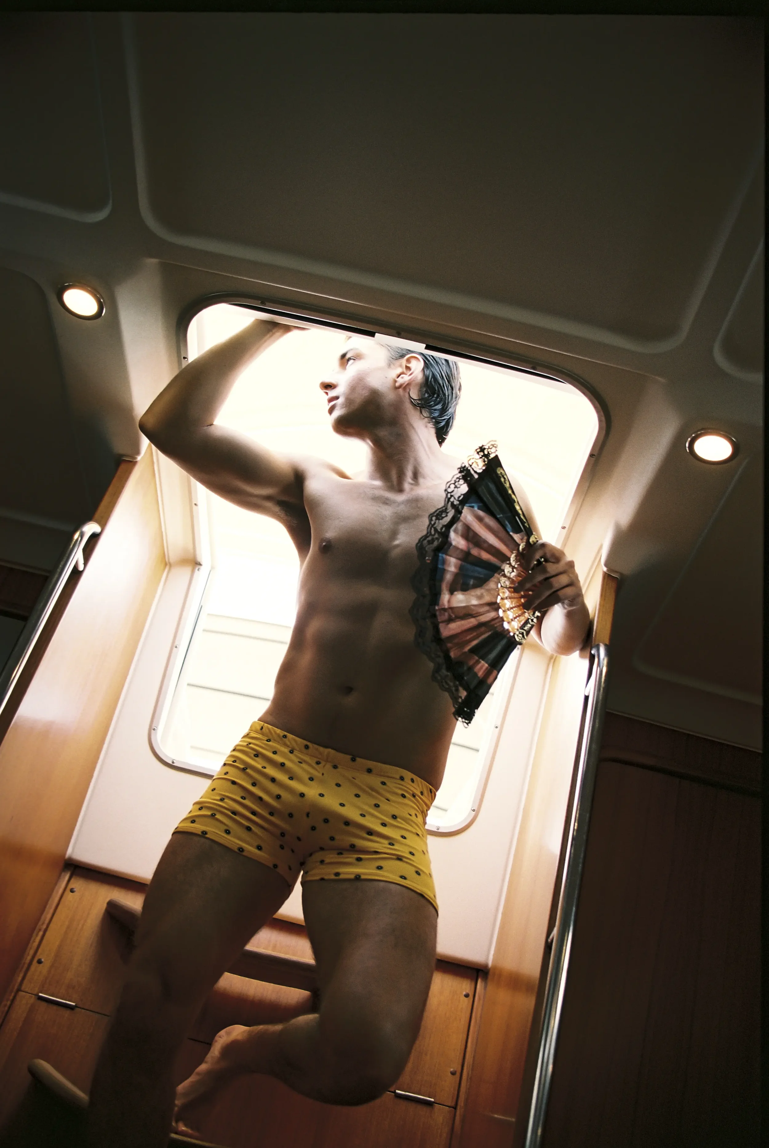 Hombre de pie, sin camisa, usando pantalones amarillos con puntos negros, sosteniendo un abanico, en el interior de una embarcación, frente a una ventana que muestra el cielo claro.
