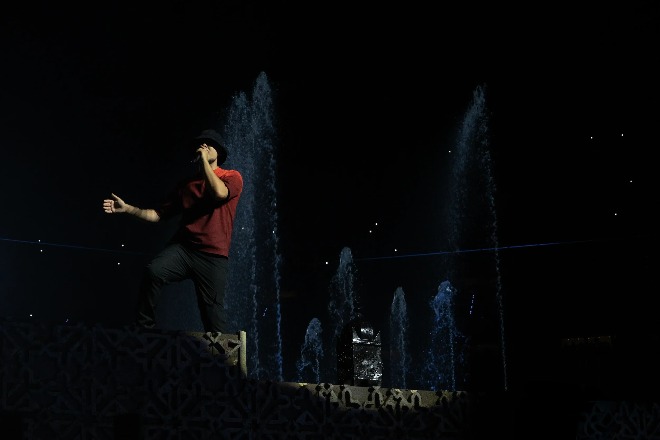 Persona con sombrero negro y camiseta roja en un escenario oscuro, frente a fuentes de agua iluminadas y estrellas en el cielo.