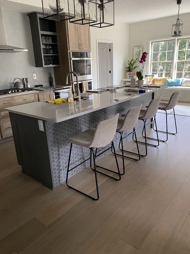 Modern kitchen with island, four beige barstools, wooden cabinets, gray tile, and a large window with colorful cushions