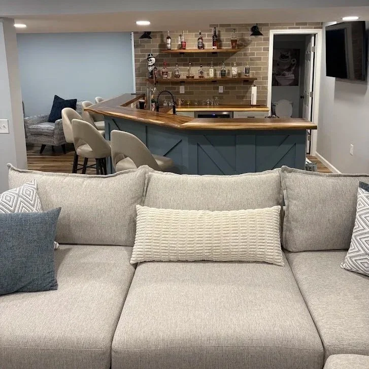 Living room with a beige sofa and decorative pillows in front of a kitchen with a blue bar counter, open shelving with bottles, and a brick backsplash.