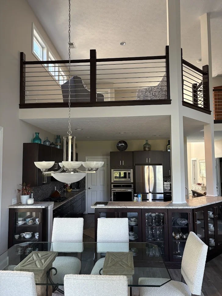 Interior of a modern home with an open kitchen and dining area, featuring a glass-top table with six upholstered chairs, dark wood cabinets, stainless steel appliances, a granite countertop, and a loft with dark wood railing.