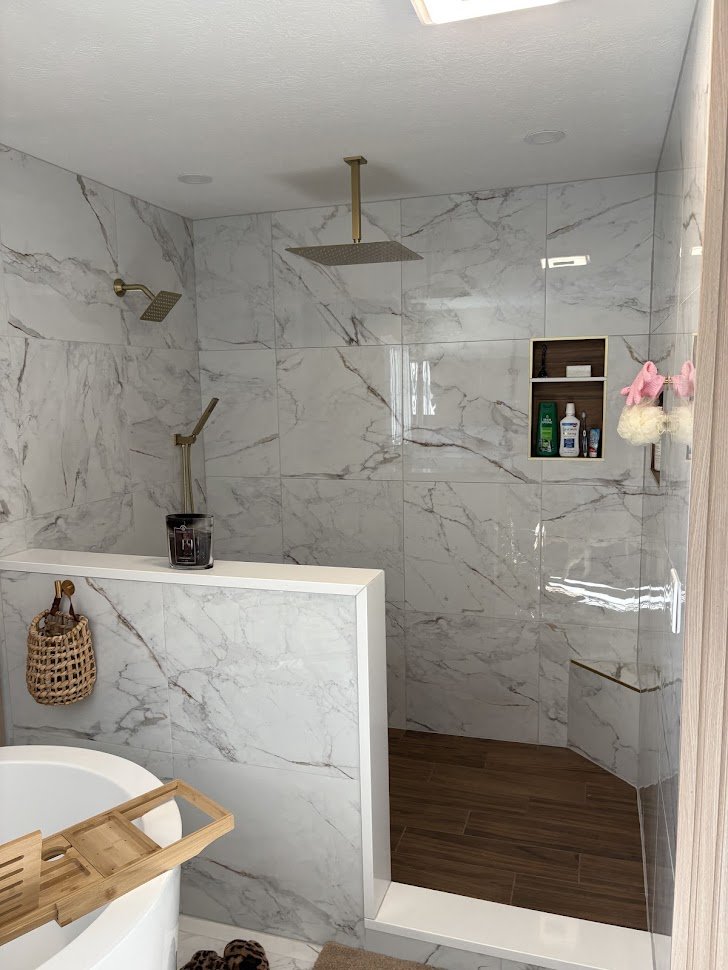 Modern walk-in shower with marble tiles, a rainfall showerhead, a handheld shower, built-in shelf with toiletries, and a wood-finish floor section.