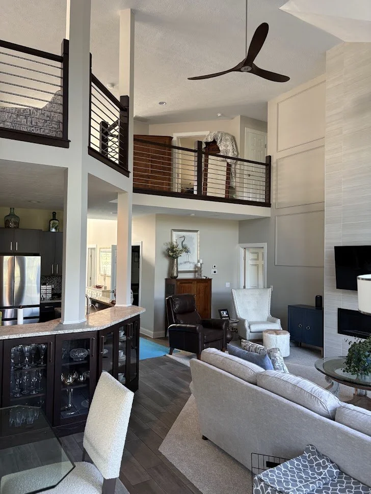 Open-concept living room with high ceiling, ceiling fan, and two-story balcony with wooden railing, viewing into the living area with gray sofa, white armchair, and fireplace, with kitchen in background.