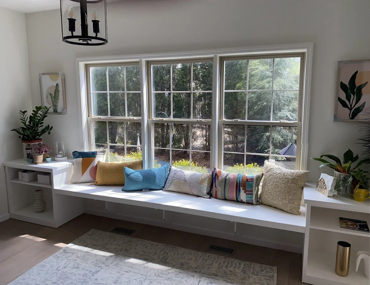 Sunlit window seat with decorative pillows, plants, and artwork in a cozy living room.