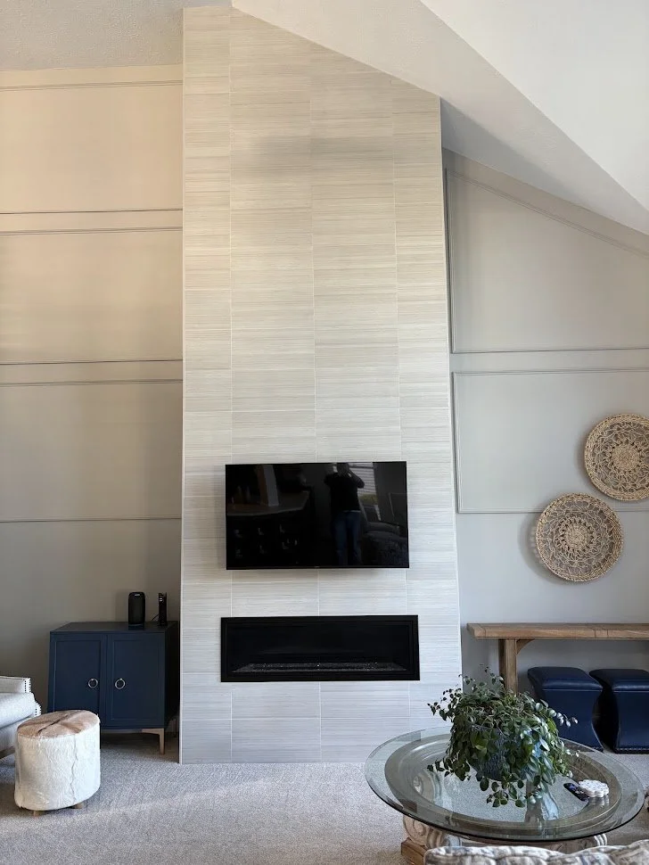 Living room with a modern fireplace and wall-mounted TV on a white textured wall, with decorative wicker wall hangings and a glass-topped coffee table with greenery.