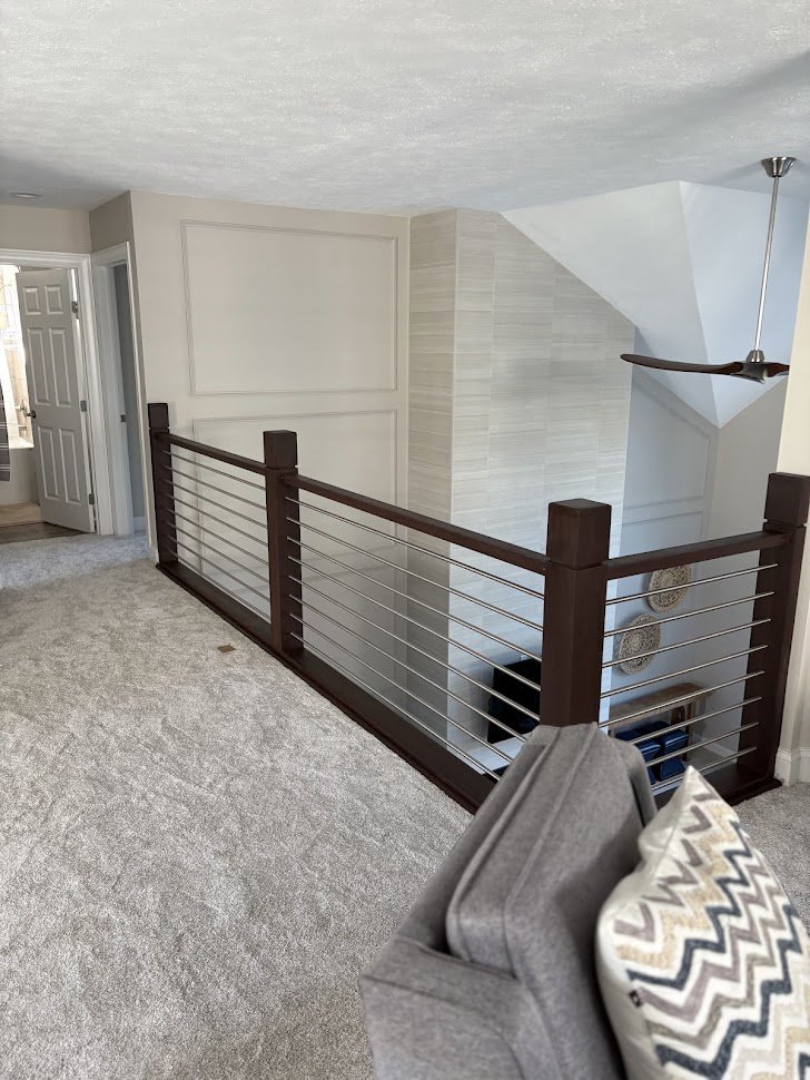 Interior view of a living space with a gray carpet, a gray armchair with a patterned pillow, a wooden railing with metal wires, a ceiling fan, and an open door leading outside.