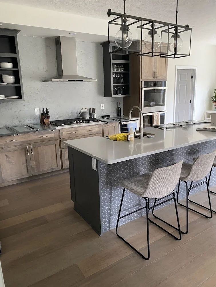 Modern kitchen with wooden cabinets, stainless steel appliances, an island with a white countertop, two beige chairs, and a black hanging light fixture with exposed bulbs.