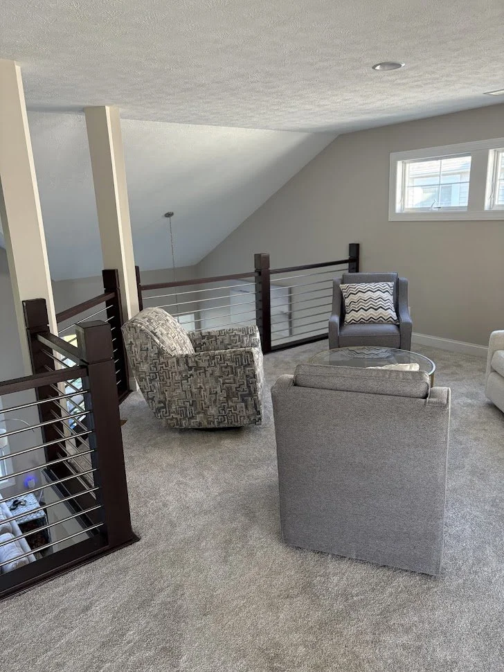 A cozy loft area with gray carpet, gray armchairs, a small round glass table, and a black railing with horizontal metal bars. A small window lets in natural light.