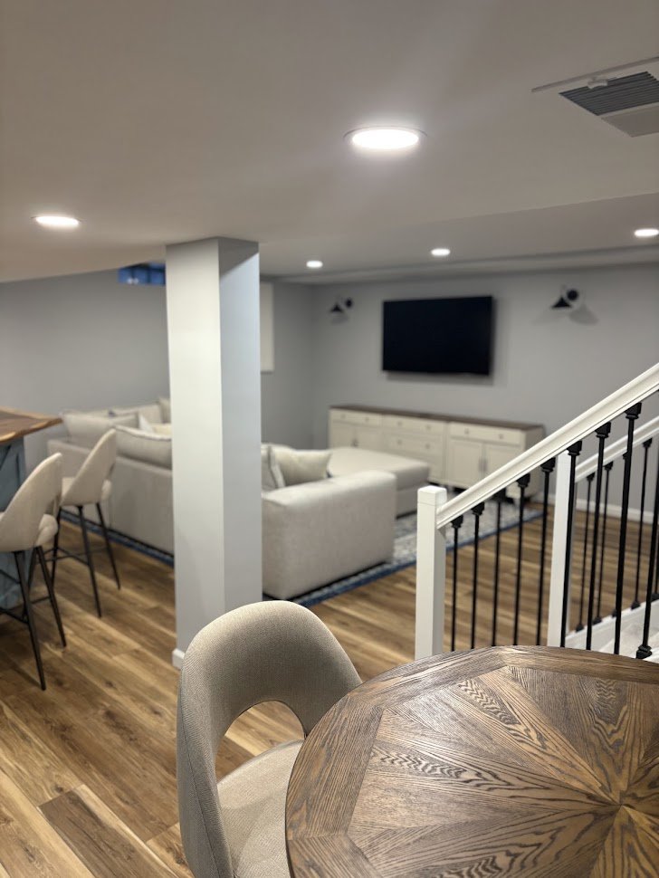 A spacious basement living area with a flat-screen TV mounted on the wall, a white sofa set, and a wooden dining table with chairs. The room has wood flooring, recessed ceiling lights, and a staircase with a black railing.