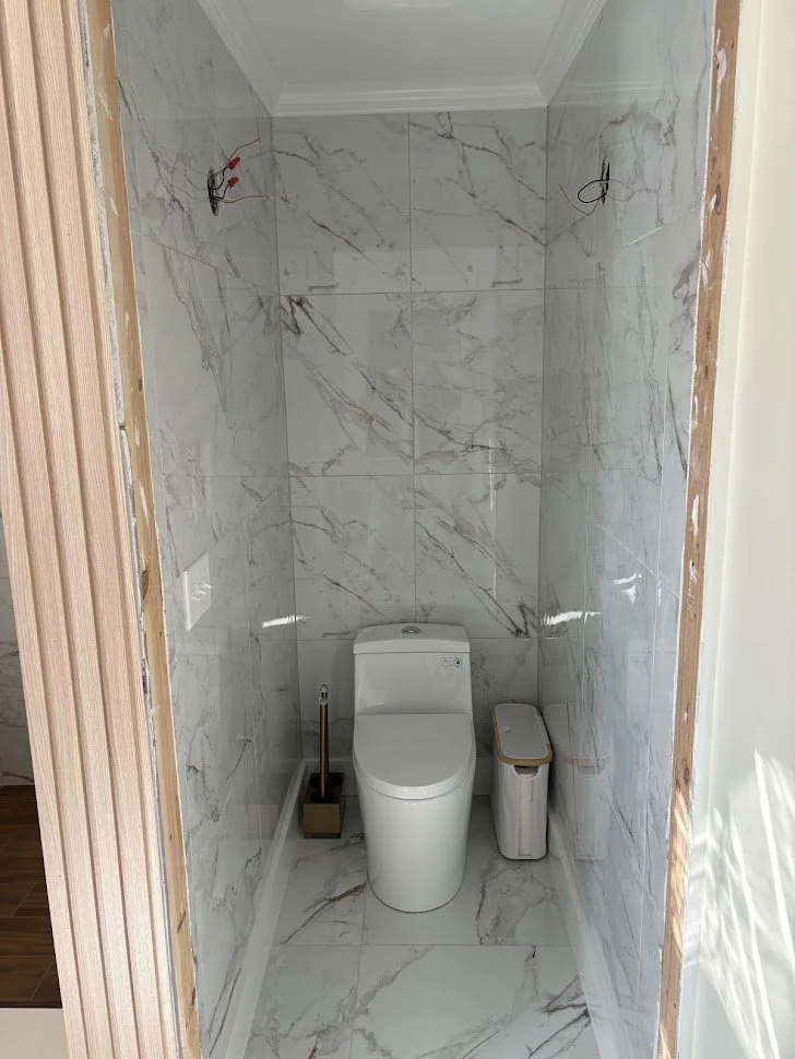 Bathroom with large white marble tiles, toilet, toilet brush, and small trash bin. Wiring on the wall indicates ongoing construction or renovation.