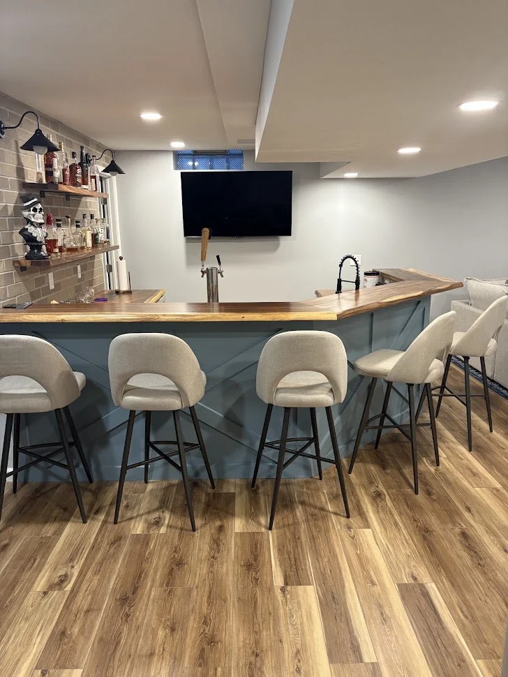 Home bar with six beige bar stools, a wooden countertop, a mounted flat-screen TV, a black sink faucet, and shelves with bottles and decorations.