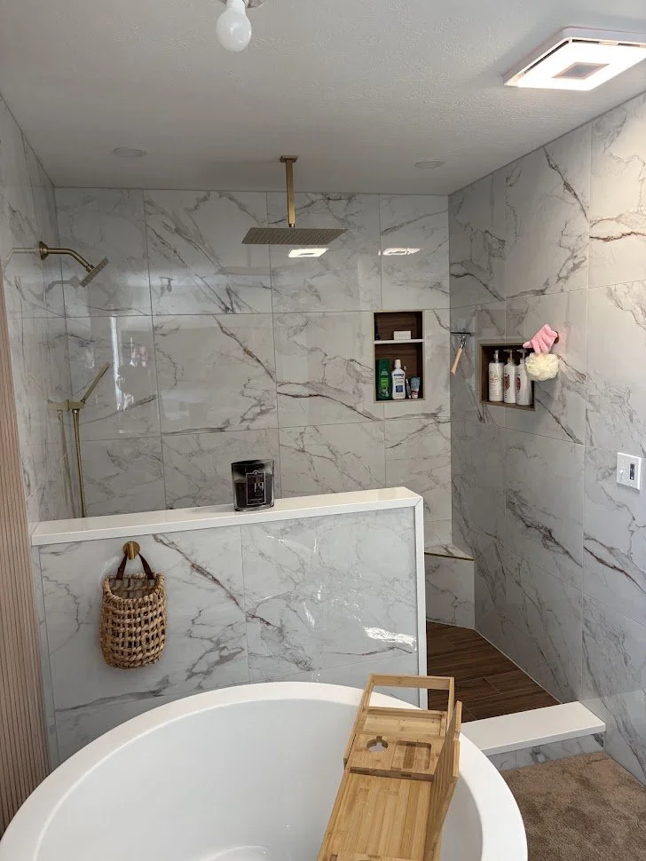 Bathroom with marble tile walls, a rainfall showerhead, a separate handheld shower, and a white bathtub with a wooden bath tray. There are built-in wall shelves holding toiletries, a woven basket hanging on a hook, and a wooden shower bench.