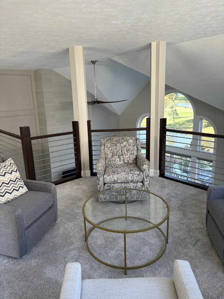 Living room area with a beige carpet, an armchair with a geometric pattern, and a glass-top round coffee table. Two gray sofas and a decorative pillow with a chevron pattern are partially visible. There is a railing with dark wood and metal wires, overlooking a space with large, arched windows showing a green landscape outside. The ceiling has sloped angles, a ceiling fan, and a unique triangular design in the background.