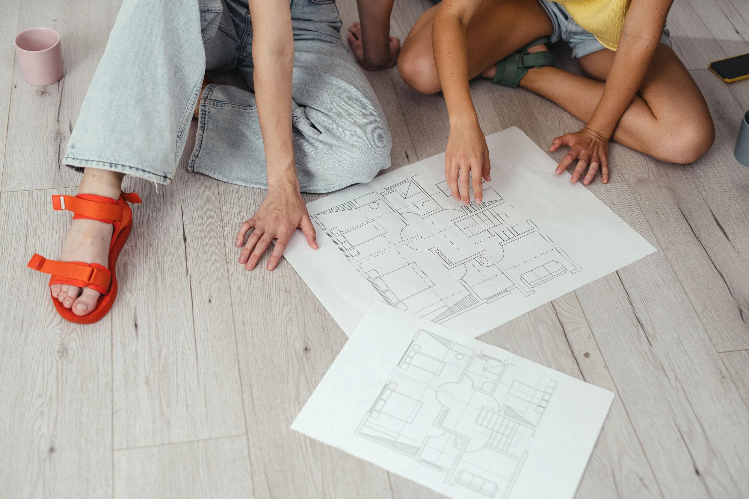 Two children sitting on the floor with architectural blueprints and drawings, one wearing light blue jeans and orange sandals, the other wearing shorts and a yellow top.