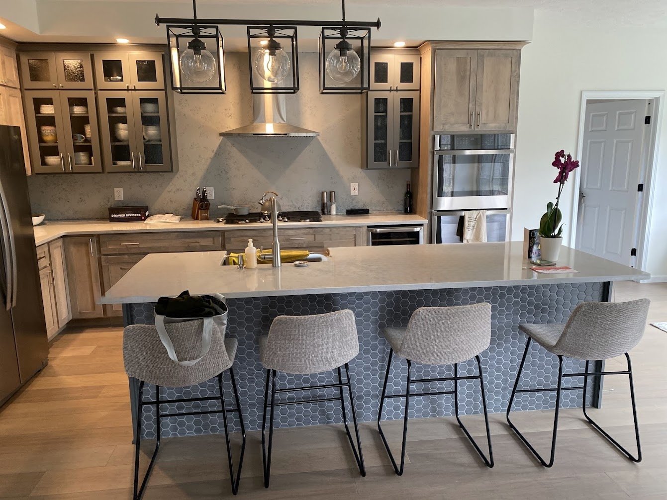 Modern kitchen with a large central island, four chairs, wooden cabinetry, stainless steel appliances, and a pendant lighting fixture with four glass bulbs.