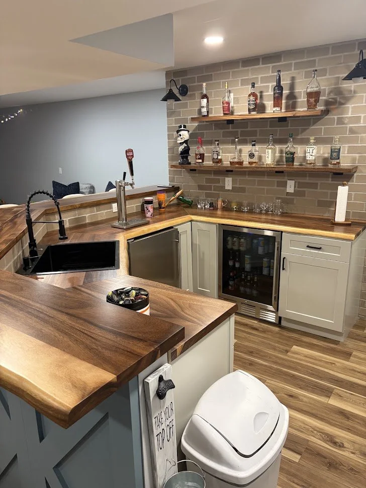 A kitchen with a wooden bar counter, a black sink, and a small refrigerator. There are two floating shelves with liquor bottles, a liquor bottle-shaped lamp, and a panda bear figure. The countertop has a paper towel roll and various small items. The floor is wood, and the wall features a brick backsplash. A sign on the trash bin says 'Take your top off'.