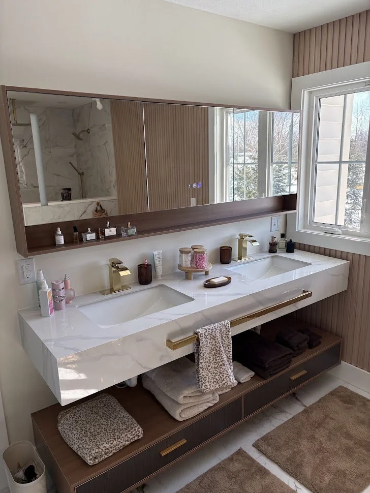 A modern bathroom vanity with dual sinks, gold faucets, a large mirror, and various toiletries, with a window revealing a snowy outdoor scene.