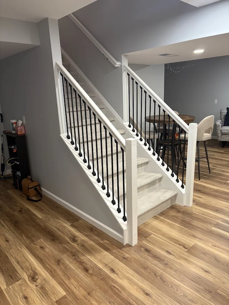 Interior view of a staircase with beige carpeted steps, white risers, and black metal spindles, leading upstairs in a home with wood flooring and gray walls.