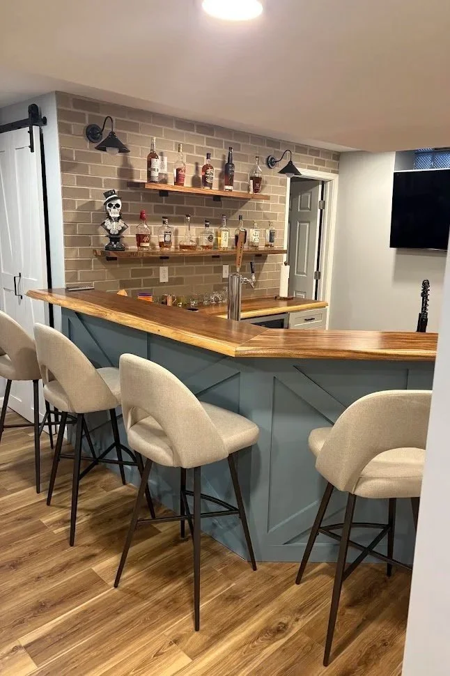 Home bar with beige bar stools, wooden countertop, brick wall, shelves with various liquor bottles, skeleton figurine, and a TV on the white wall.