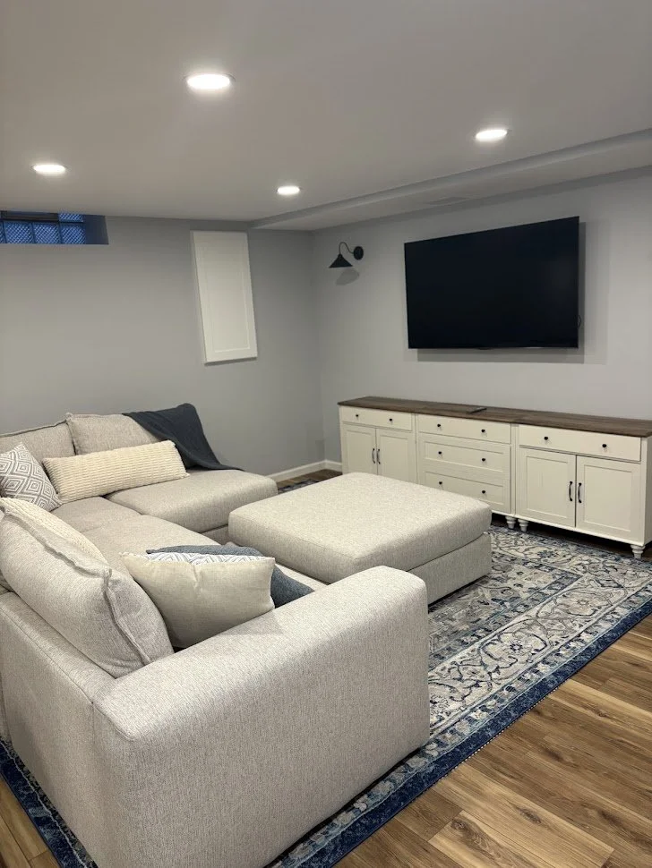 Living room with beige sectional sofa, throw pillows, a patterned area rug, a white cabinet, a wall-mounted flat-screen TV, and wall-mounted light fixtures.