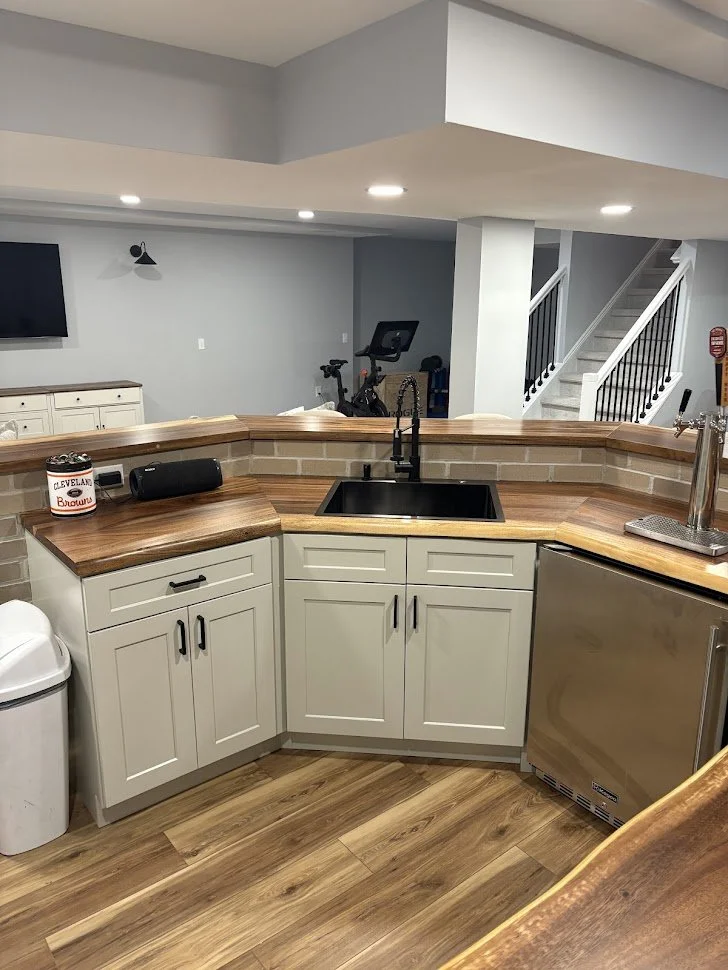 Kitchen with white cabinets, wooden countertops, a black sink, and a small refrigerator. There is a open space in the background with a TV, exercise bike, and stairs leading upstairs.