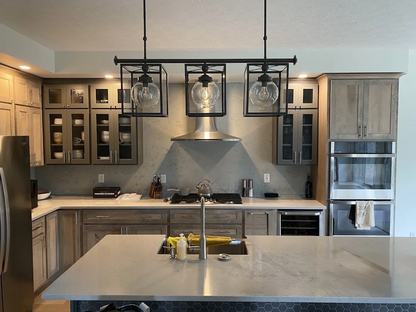 Modern kitchen with wood cabinets, a central island with a sink, pendant lighting, stainless steel appliances, and a backsplash with a light textured finish.