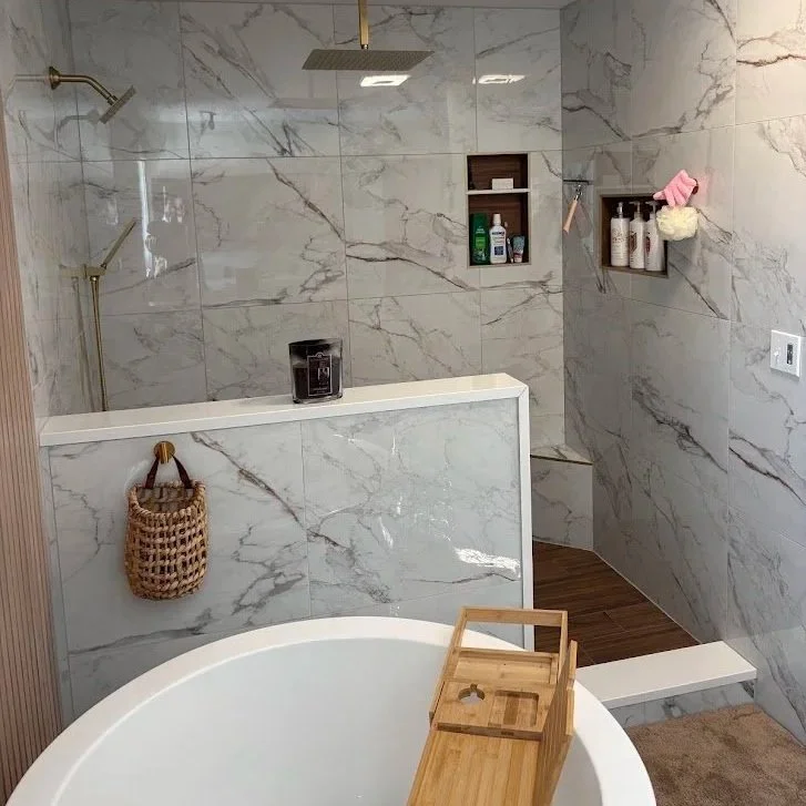 Bathroom with marble walls, a bathtub with a wooden tray, a shower with gold fixtures, and wall-mounted shelves holding toiletries and a pink towel.