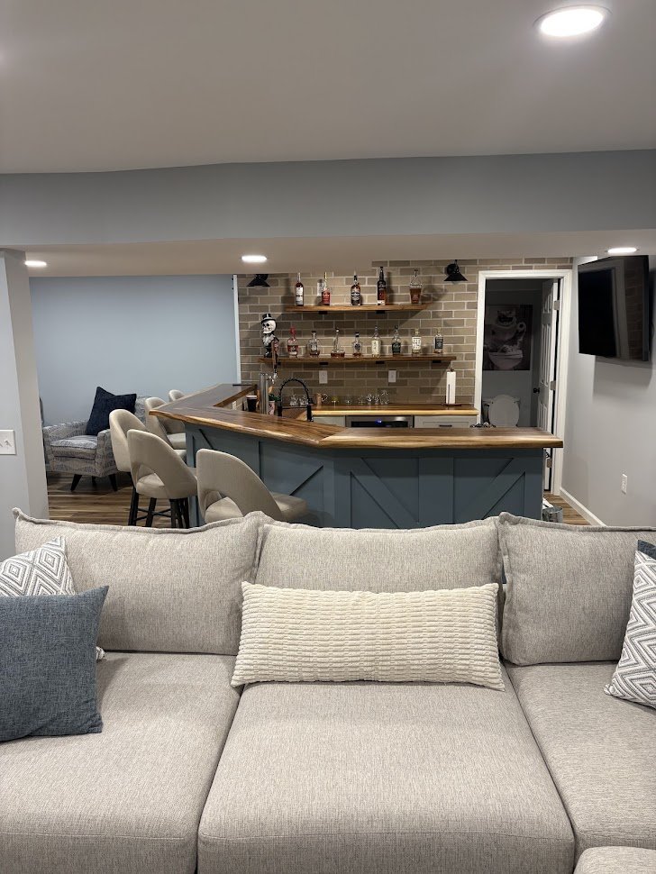 A living room with a gray sofa and various pillows, a bar area with a wooden counter and beige barstools, a brick wall with shelves holding liquor bottles, and a nearby seating area with a patterned chair.