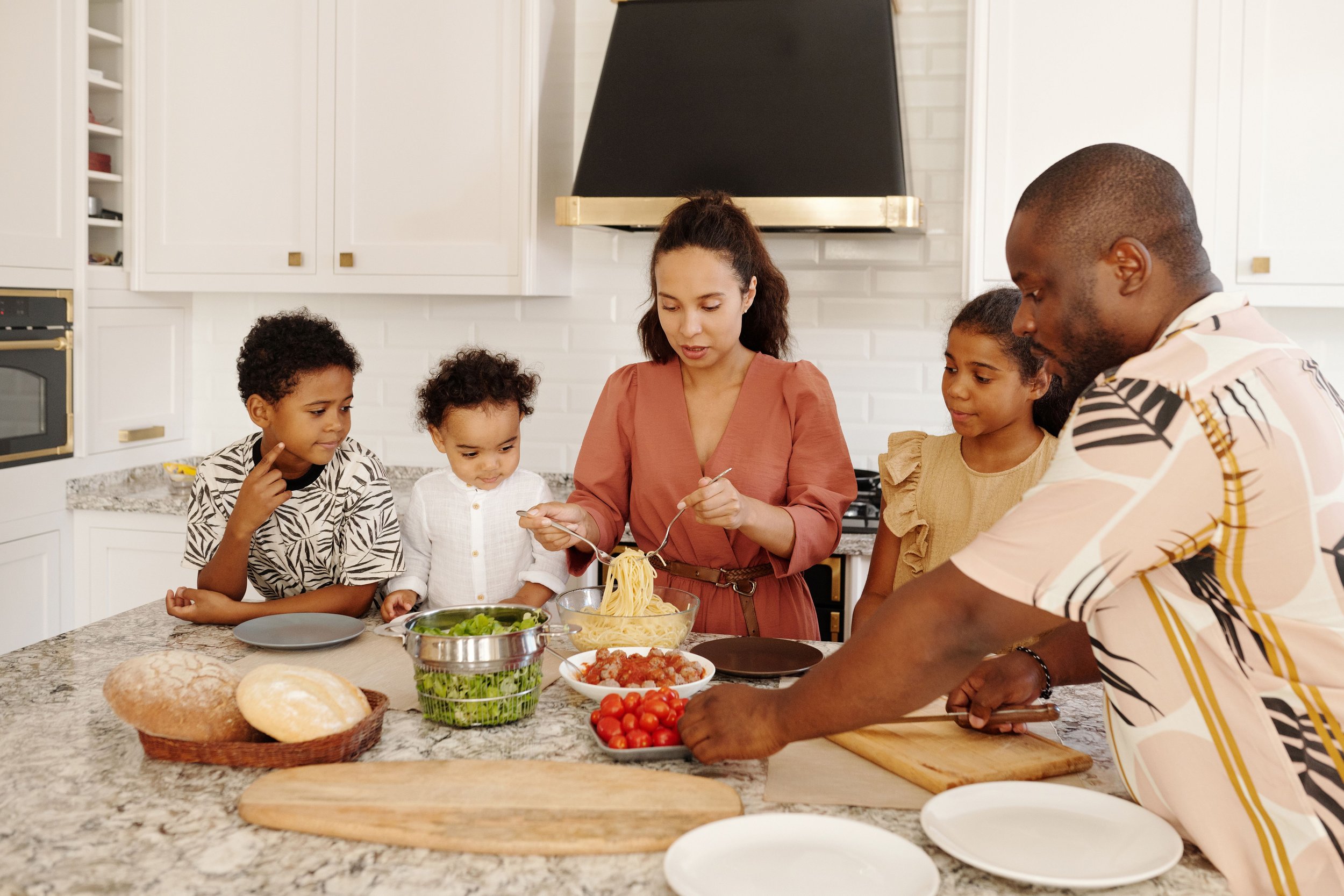 A family preparing a meal together in a kitchen, with vegetables, bread, and pasta on the counter.