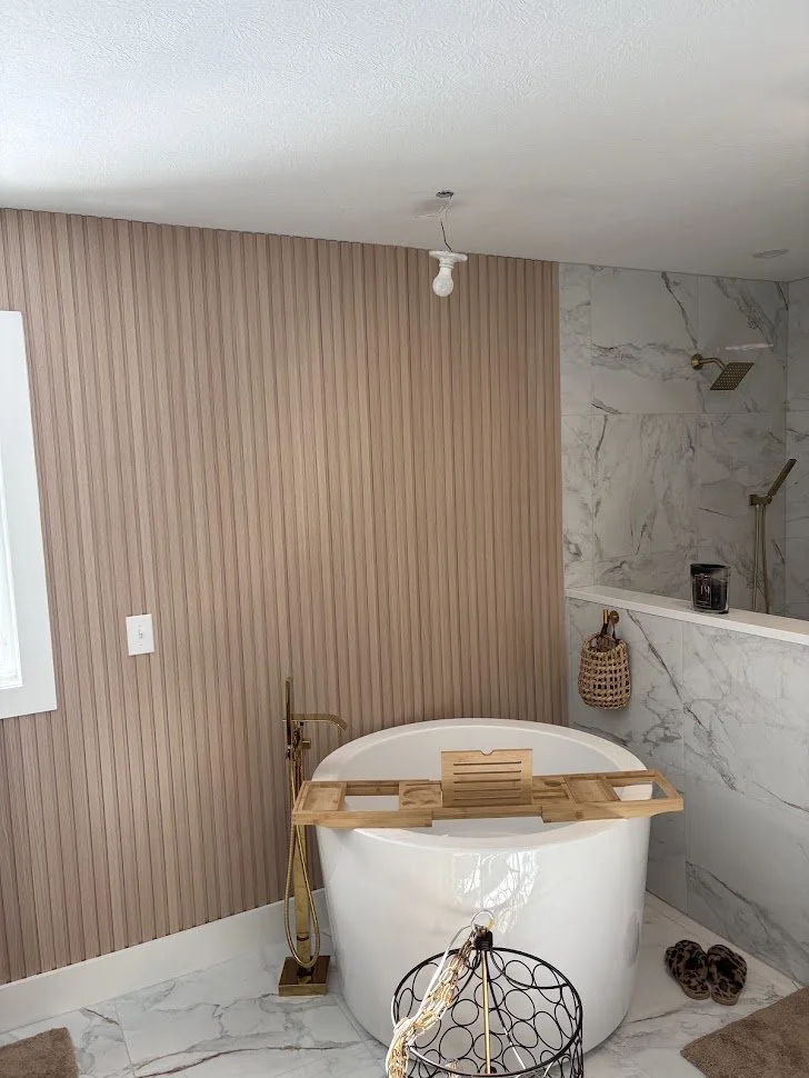 Bathroom with a freestanding bathtub, bamboo bath tray, gold faucet, marble tiled shower area, hanging basket, slippers, light bulb hanging from ceiling, and a wall with vertical beige paneling.