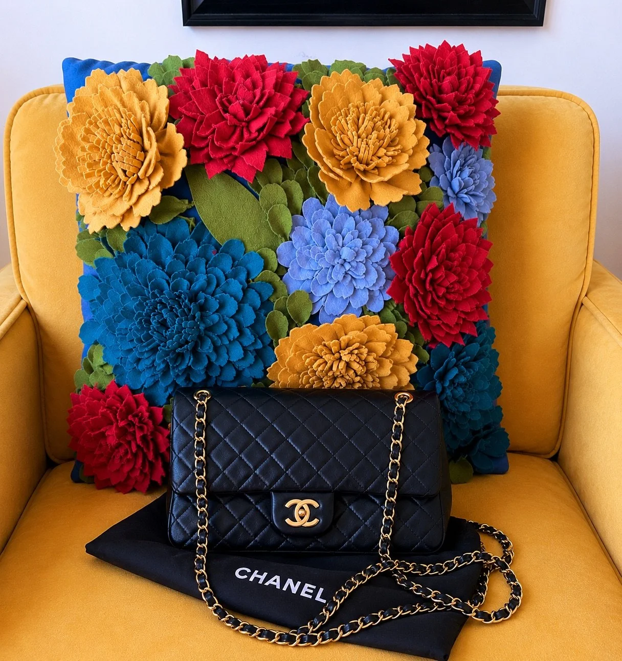 A black quilted Chanel purse with a gold chain strap and CC logo on the front, placed on a black Chanel dust bag, in front of a colorful floral cushion with large yellow, blue, red, and purple flowers, on a yellow armchair.