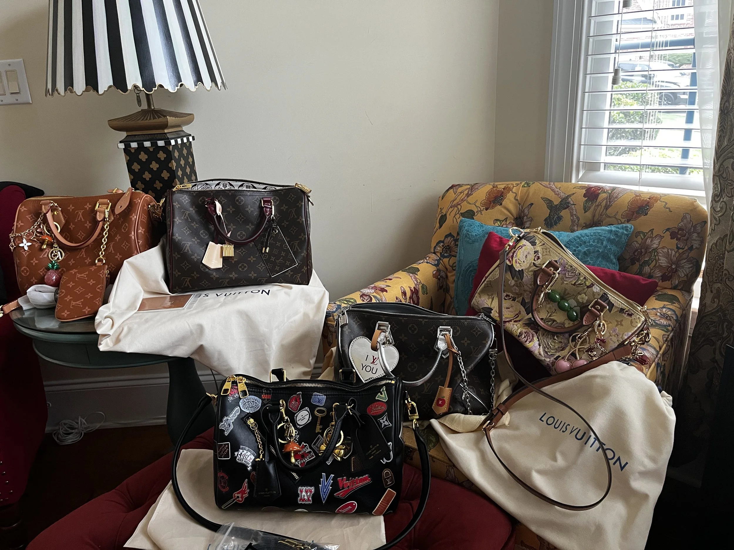A table displays various designer handbags, with some bags placed on white dust bags. There is a floral patterned couch with colorful pillows in the background and a window with blinds letting in natural light. A lamp with a black and white striped shade is on the wall.