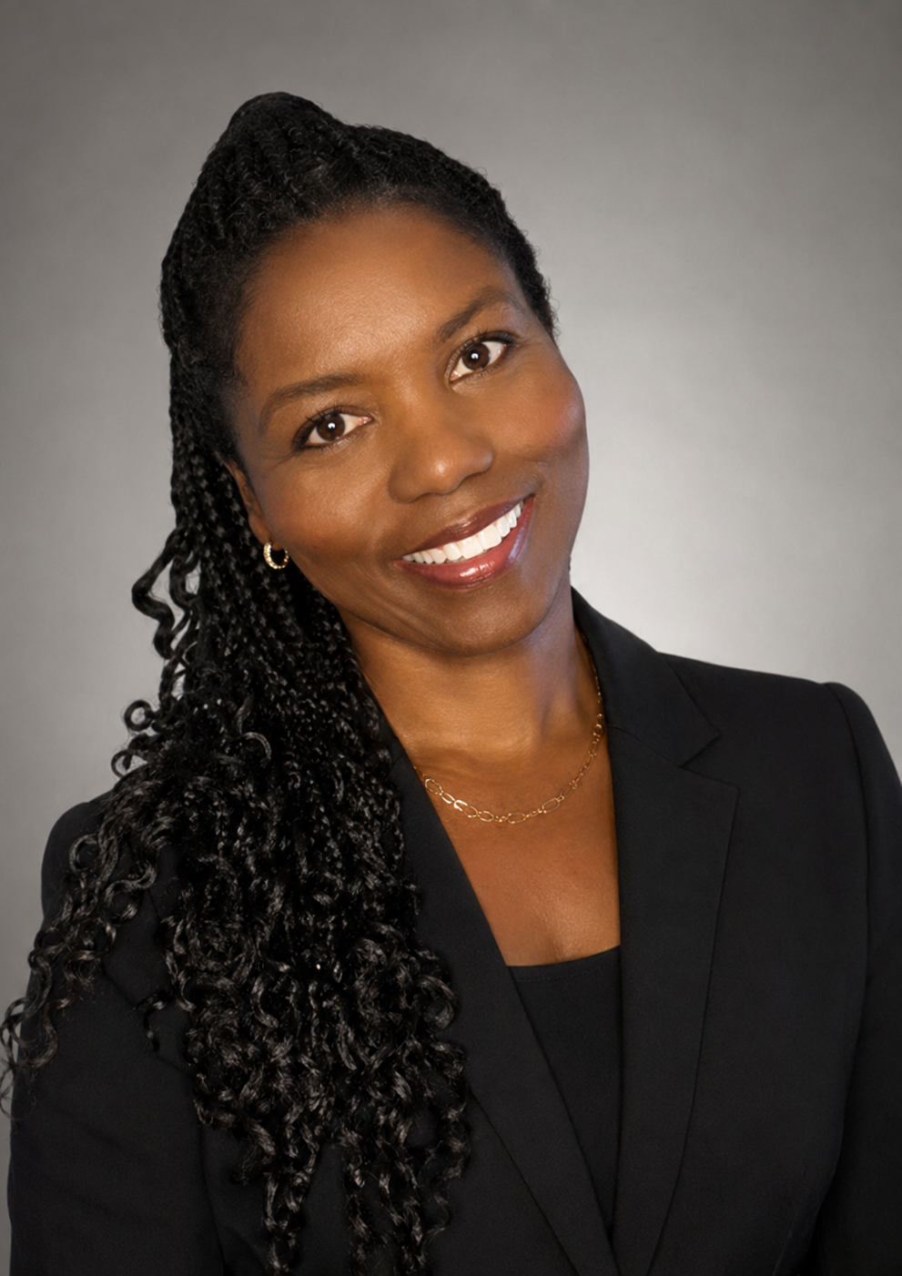 A professional woman with braids, wearing a black blazer and gold jewelry, smiling at the camera against a gray background.